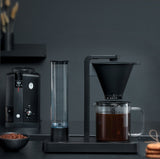 A PERFORMANCE BLACK coffee maker with a transparent water reservoir and a glass carafe filled with coffee. Featuring unique technology for the perfect temperature, it boasts two award badges: one with a laurel wreath indicating "Best i Test Tek.no" and another that reads "Testvinner" with stars and a ribbon symbol.